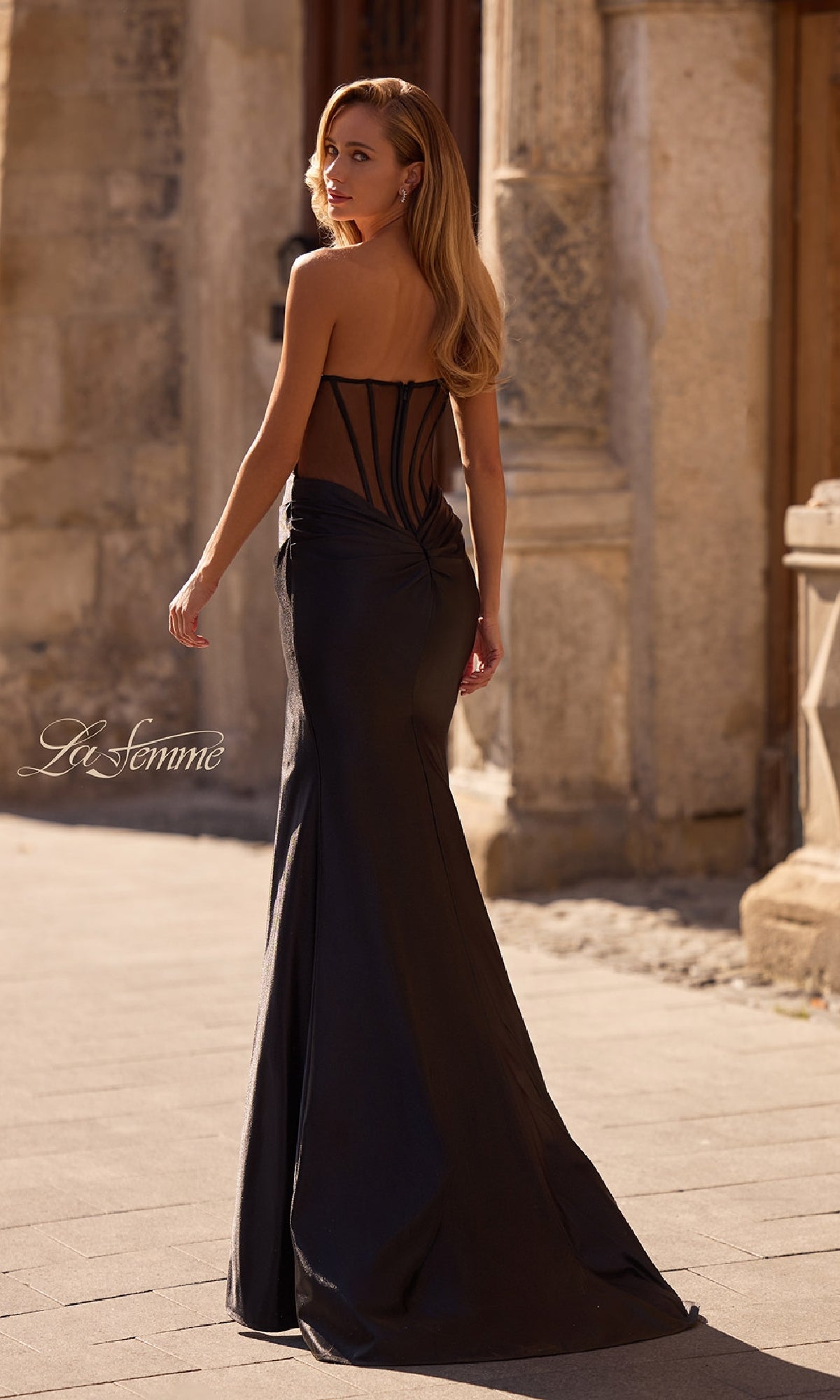 A woman in the Sheer-Back Strapless Prom Gown: La Femme 32649, featuring a sweetheart neckline, fitted silhouette, and sheer back panel, stands on a stone walkway outdoors and looks over her shoulder.